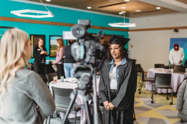 A woman in graduation cap and gown standing next to a camera.