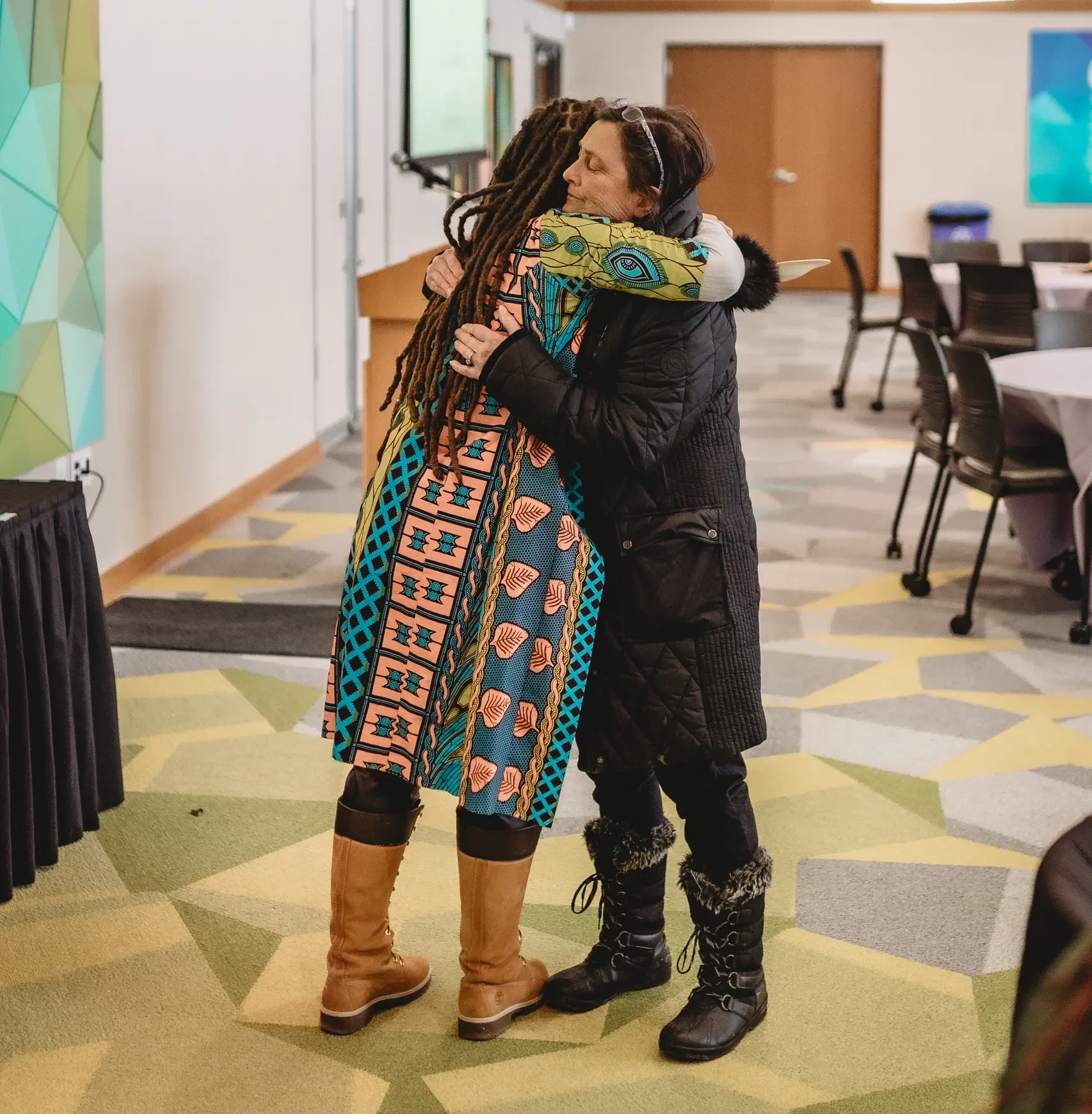 Two people hug in a room with chairs.