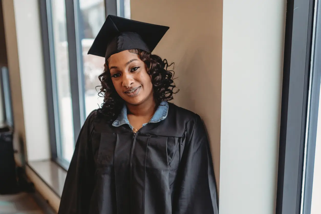 A woman in graduation gown and cap standing next to wall.